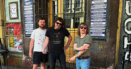 Das Bild zeigt die drei Mitglieder:inenn der Band Trigger Kid. Sie stehen in sommerlicher Kleidung vor einer Hausfassade mit Graffiti und Plakaten und schauen in die Kamera. Zwei der Personen tragen eine Sonnenbrille.