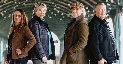 Das Bandfoto zeigt die 4 Mitglieder:innen der Band Bornland. Jeweils 2 Personen stehen Rücken an Rücken und schauen lächelnd in die Kamera. Im Hintergrund ist eine runde Überdachung mit Lichterketten zu erkennen. Vermutlich wurde das Bild am Gleis des Kassler Hauptbahnhofs aufgenommen.