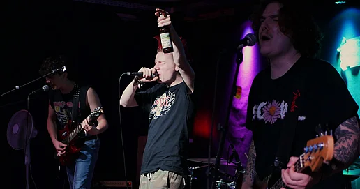 Das Bild zeigt die Band Bierkenstock während eines Konzerts. Mittig und am hälsten angestrahlt sieht man den Sänger der Band mit einem Mikrofon und einer Flasche Bier in der Hand. Am linken Bildrand erkennt man eine Person, die Gitarre spielt und gleichzeitig in ein Mikrofon singt. Am rechten Bildrand spielt eine Person Bass und singt ebenfalls in ein Mikrofon.