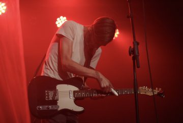 Ein Mensch steht in einer gekrümmten Haltung mit einer Gitarre auf einer Bühne. Er wird von hinten mit licht agestrahlt, so dass man sein Gesicht kaum erkennen kann. Statt die Gitarre so zu spielen, wie es üblich ist, hält der Mensch einen kleinen weißen Gegenstand gegen die Saiten und erzeugt vermutlich mit diesem die Klänge.