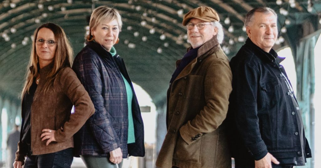 Das Bandfoto zeigt die 4 Mitglieder:innen der Band Bornland. Jeweils 2 Personen stehen Rücken an Rücken und schauen lächelnd in die Kamera. Im Hintergrund ist eine runde Überdachung mit Lichterketten zu erkennen. Vermutlich wurde das Bild am Gleis des Kassler Hauptbahnhofs aufgenommen.